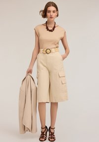 Beige ribbed top, beige cargo shorts with pockets, woven belt with round buckle, holding a beige jacket, and wearing strappy brown heels.