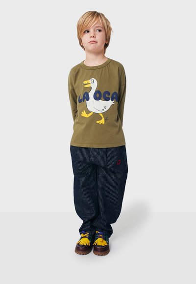 Grünes Langarmshirt mit einem Entenmotiv und dem Text "LA OCA", kombiniert mit blauen gestreiften Hosen und braunen Schuhen mit gelben Akzenten.