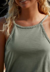 Top de tirantes sin mangas en verde oliva con un cuello trenzado. La tela parece suave y lisa, presenta un corte holgado y costuras mínimas.