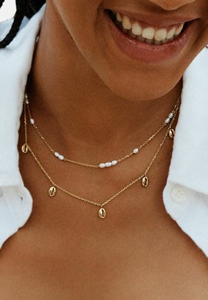 Osoba koja se smiješi i nosi slojevite zlatne ogrlice s malim bijelim biserima i zlatnim privjescima u obliku zrna kave, odjevena u bijelu košulju s ovratnikom.