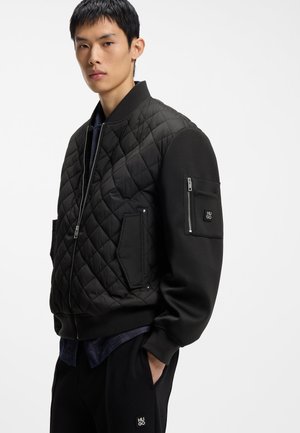 Jeune homme portant une veste bomber matelassée noire avec des poches zippées et le logo HUGO sur la poche de la manche et le pantalon.