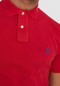 Czerwona koszulka polo z teksturowanej tkaniny, z niebieskim logo na lewej piersi, standardowym kołnierzykiem oraz zapięciem na dwa guziki.