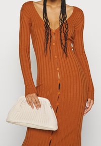 Robe côtelée couleur rouille avec un col en V profond, manches longues et boutons sur le devant. Pochette tissée blanche tenue dans la main droite.