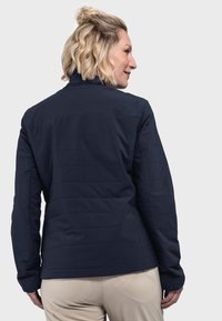 Frau mit kurzem, welligem, blonden Haar, die eine dunkelblaue, gesteppte Jacke und beigefarbene Hosen trägt und von der Kamera weggedreht ist, vor einem schlichten, hellen Hintergrund.