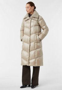 Beige pufferjack met een grote, getextureerde kraag; chevronpatroon; knielange stijl; zijzakken; gedragen over donkere, wijde broekspijpen en zwarte laarzen.