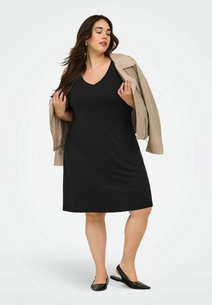 Kvinde iført en sort ærmeløs kjole, beige jakke lagt over skuldrene, sorte slingback-sko, står mod en ensfarvet hvid baggrund.
