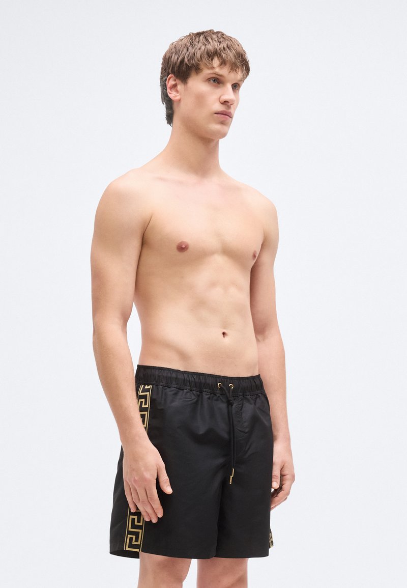 Shorts de bain noirs en matériau léger, présentant un motif géométrique doré sur les côtés et une taille élastique avec cordon de serrage.