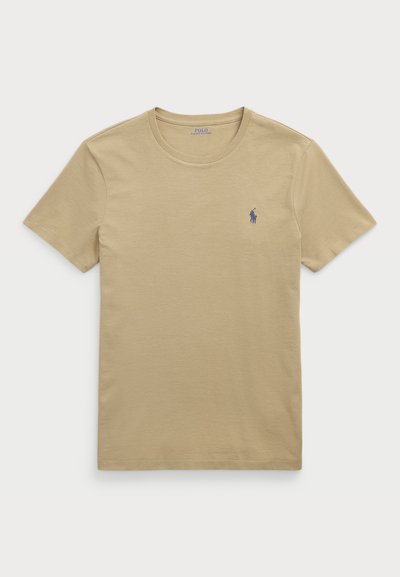 Beżowa bawełniana koszulka z klasycznym okrągłym dekoltem, krótkimi rękawami i niebieskim logo po lewej stronie klatki piersiowej. Gładka faktura.