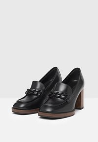 Mocassins à talons en cuir noir avec un accent de chaîne épaisse. Présente un bout arrondi et un talon en bois avec une finition texturée.