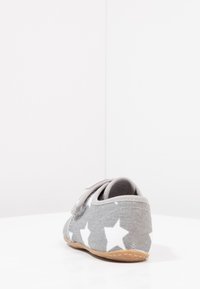 Chaussure en tissu gris avec de grands motifs d'étoiles blanches, une sangle Velcro, une forme arrondie et une semelle en caoutchouc flexible.