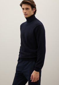 Suéter de cuello alto azul marino hecho de tela suave, con un diseño ajustado y puños acanalados, combinado con pantalones oscuros.