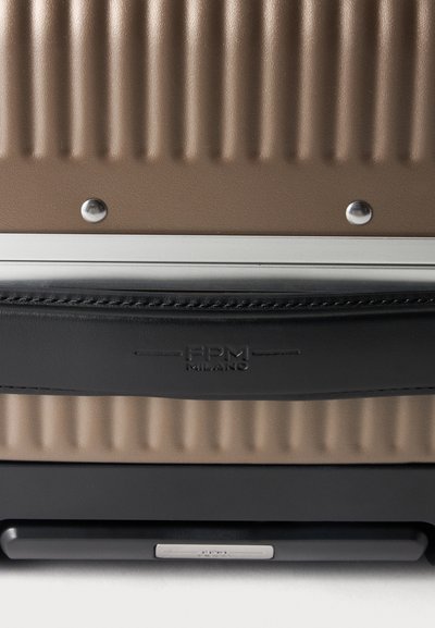 Gros plan d'une valise marron côtelée avec une poignée en cuir noir embossée BPM Milano et des rivets métalliques assurant une bordure argentée.