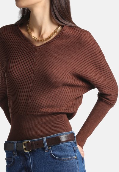 Femme portant un pull côtelé brun ajusté, une ceinture marron foncé, un jean bleu et un collier chaîne doré, debout avec une main sur la hanche.