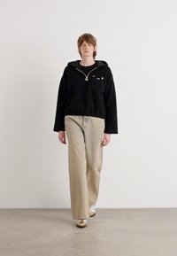 Pulôver de fleeces pretos com colarinho com fecho e bolso no peito, combinado com calças de perna larga em bege claro e sapatos metálicos. Fundo neutro.