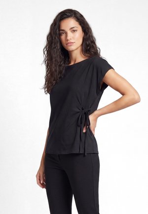 Femme aux longs cheveux ondulés portant un haut noir à manches courtes avec détail nœud sur le côté et un pantalon noir, posant une main sur la hanche.