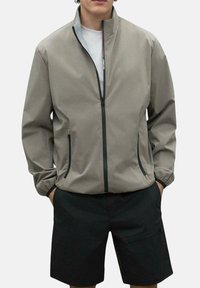 Chaqueta ligera beige con un alto cuello, cremallera frontal y bolsillos laterales; cuenta con puños elásticos y una textura suave. Combinada con pantalones cortos negros.