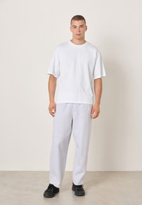 Bela bombažna majica ohlapnega kroja, kombinirana z sivimi športnimi hlačami. Robustni črni športni copati. Enostavna podlaga poudari pozornost na oblačilo.