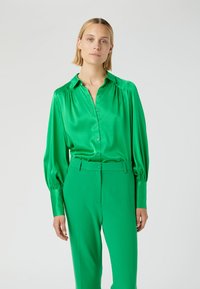 Groene satijnen blouse met een kraag, knopen aan de voorkant en opgeblazen lange mouwen, gecombineerd met bijpassende groene maatwerk broek.