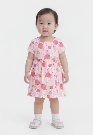 Παιδάκι που στέκεται φορώντας ένα ροζ φόρεμα με σχέδιο φράουλες, λεμόνια, λουλούδια και λευκά σανδάλια με κάλτσες σε απλό φόντο.