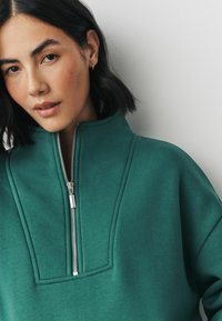 Grøn sweatshirt lavet af struktureret stof, med en høj krave, metallynlås og syede detaljer langs skuldrene.