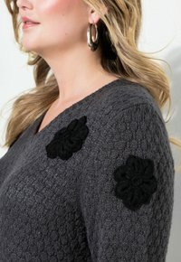 Pull en tricot gris avec un col en V, tissu texturé et appliqués floraux noirs sur les épaules. Grandes boucles d'oreilles en argent visibles.