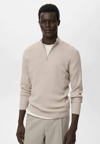 Suéter de punto acanalado beige con cuello de media cremallera, mangas largas y tela texturizada. Se lleva sobre una camisa de color crema claro, combinado con pantalones claros.