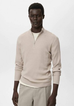 Beige ribbed knit sweater with a half-zip collar, long sleeves, and textured fabric. Worn over a light cream shirt, paired with light trousers.