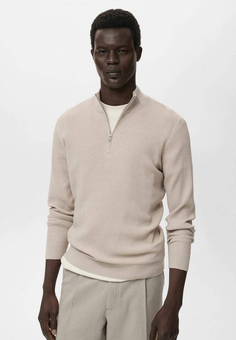 Suéter de punto acanalado beige con cuello de media cremallera, mangas largas y tela texturizada. Se lleva sobre una camisa de color crema claro, combinado con pantalones claros.