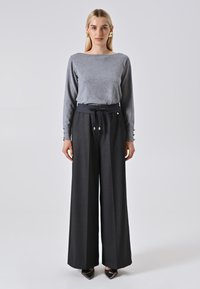 Donna in piedi con una maglietta grigia a maniche lunghe, pantaloni neri a gamba larga con vita regolabile, scarpe nere a punta e orecchini geometrici dorati.