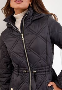 Schwarze gesteppte Jacke mit hohem Kragen, goldfarbenem Reißverschluss und verstellbaren Kordelzügen in der Taille. Aus einem glatten, leichten Material mit Seitentaschen.