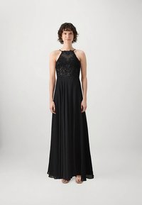 Γυναίκα με κοντά σγουρά μαλλιά, φορώντας μια αμάνικη μαύρη τουαλέτα με κορσέ από δαντέλα και ρευστή φούστα, που στέκεται μπροστά σε ένα απλό ανοιχτό φόντο.