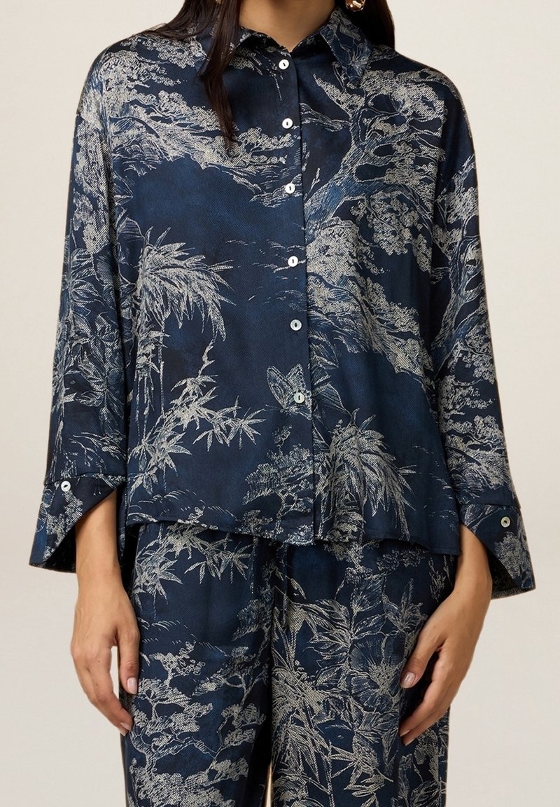 Blouse marine à coupe ample, boutonnée sur le devant, avec des manches trois-quarts, ornée de motifs botaniques argentés complexes et d'une texture lisse.