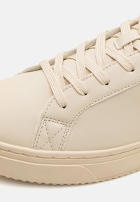 Sneaker beige in pelle con finitura texturizzata, lacci piatti e suola in gomma. Presenta cuciture sottili e design a punta rotonda.