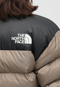 Kurtka puchowa w kolorach czarnym i taupe, z pikowanym wzorem i błyszczącym wykończeniem. Logo The North Face jest wyraźnie widoczne w białym haftowaniu.