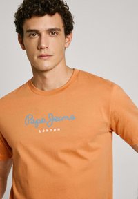 Oranžinė medvilninė marškinė su apvalia iškirpte, priekyje spausdinamu mėlynu "Pepe Jeans" logotipu ir "LONDON" tekstu. Minkšta, lygi tekstūra.