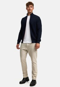Sudadera navy con cremallera y tejido texturizado, usada sobre una camiseta blanca; combinada con pantalones beige claros y botas negras con cordones.