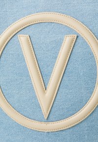 Zils denima audums ar lielu bēša ādas ovālu logotipu, kura centrā ir burts "V". Gluda tekstūra, izšūti detaļas uz logotipa.