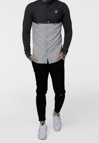 Camicia a maniche lunghe con bottoni, parte superiore in denim di colore grigio scuro e tessuto grigio più chiaro sotto. Presenta un colletto arrotondato e un logo ricamato.