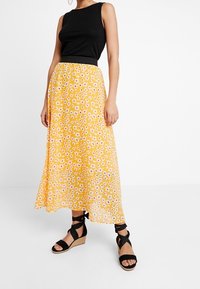 Top de tirantes negro acanalado combinado con una falda midi amarilla con flores que presenta pequeñas flores blancas y marrones. Sandalias de cuña con tiras negras.