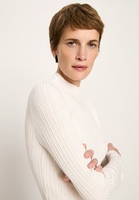 Frau mit kurzen braunen Haaren, die ein weißes, geripptes Langarmshirt trägt, steht mit verschränkten Armen da und schaut leicht zur Seite.