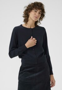 Schwarze Strickjacke mit rundem Ausschnitt, Knopfverschluss und langen Ärmeln, kombiniert mit einem strukturierten schwarzen Rock.