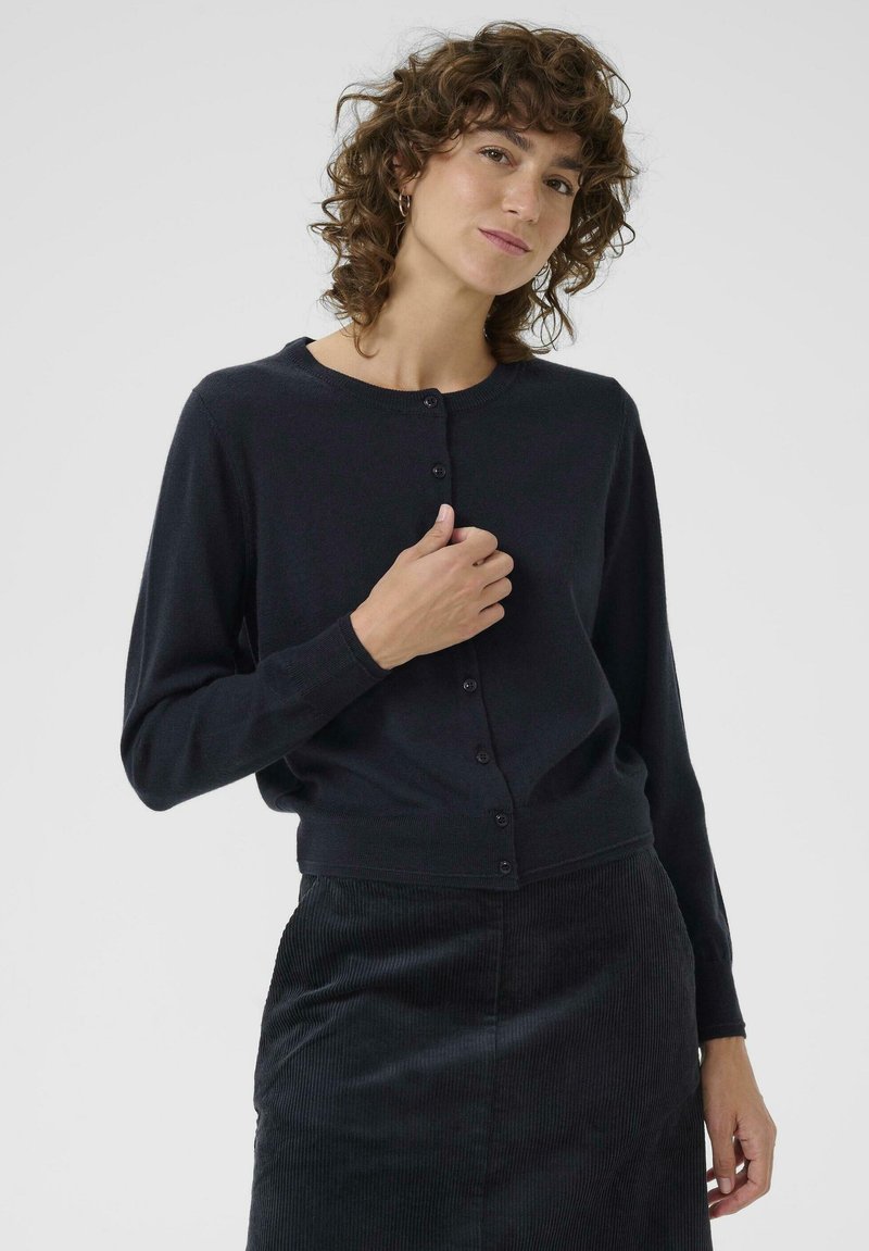 Schwarze Strickjacke mit rundem Ausschnitt, Knopfverschluss und langen Ärmeln, kombiniert mit einem strukturierten schwarzen Rock.
