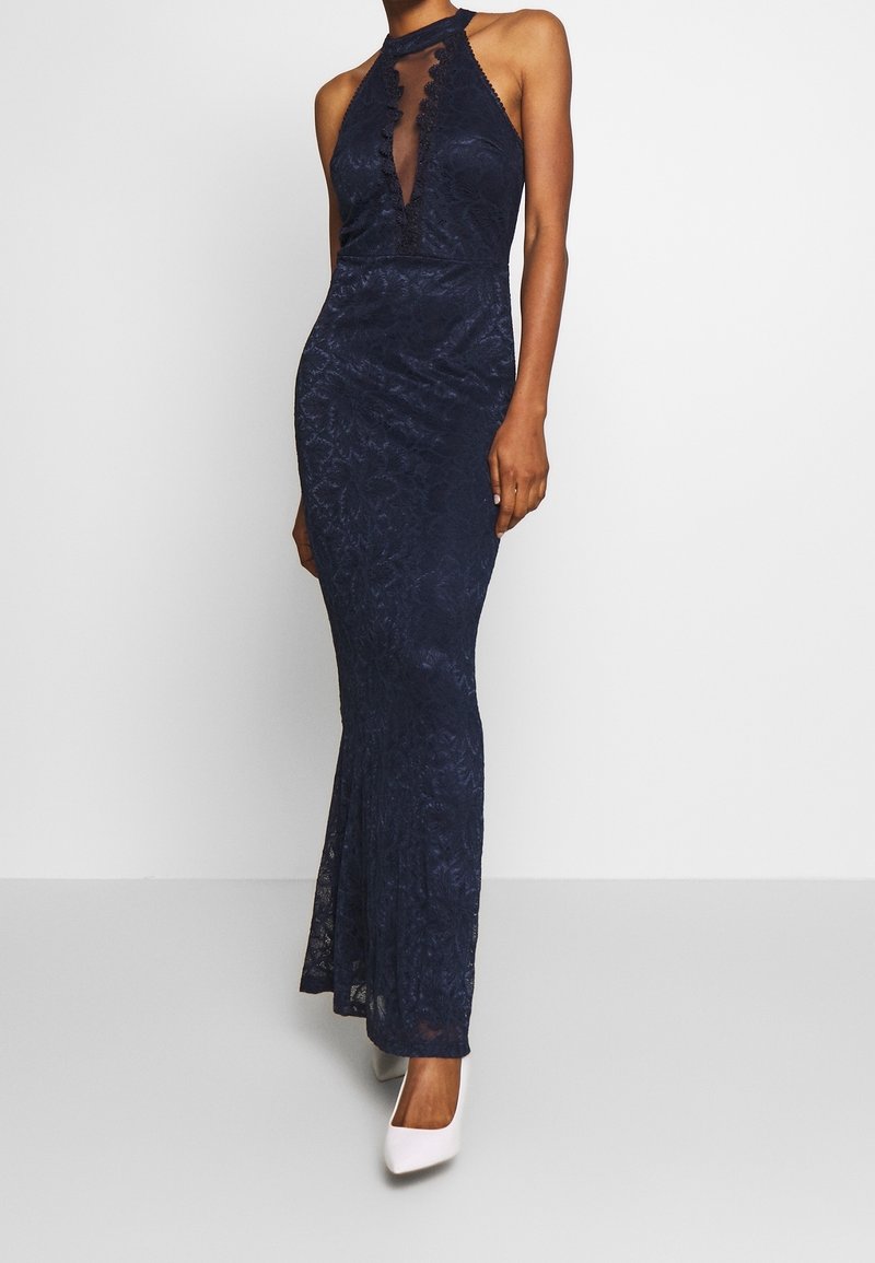 Femme portant une robe de soirée bleu marine en dentelle avec un col halter, un décolleté en V profond festonné et des talons blancs pointus.