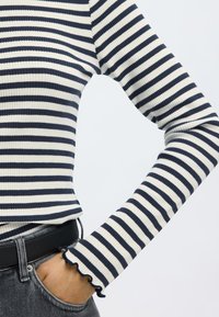 Csíkos hosszú ujjú felső tengerészkék és krémszínű bordázott anyagból, testhez simuló szabással és fodros mandzsettákkal. Szürke farmerrel és fekete övvel van stílusosan kombinálva.