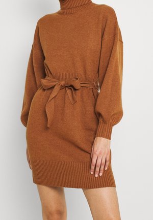Robe en maille marron à manches longues avec un col montant et une ceinture nouée à la taille, poignets et ourlet côtelés, portée par une personne debout.