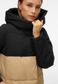 Pufjakas ar melnu augšējo daļu un bēšu apakšējo daļu. Iekļauj augstu apkakli un quilted dizainu ar gludu tekstūru.