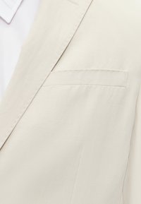 Gros plan sur le revers beige d'un blazer et la poche poitrine avec des coutures visibles au-dessus d'un col de chemise blanche.