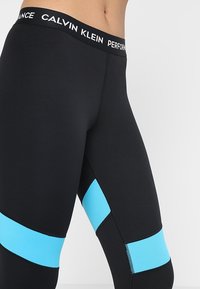 Mustat leggingit, joissa on leveä vyötäröosa valkoisella tekstillä, ja yläosassa kirkkaat siniset paneelit. Valmistettu joustavasta kankaasta.