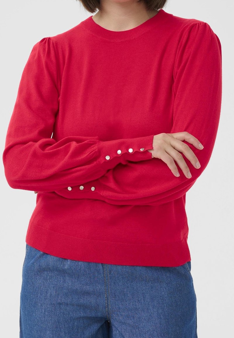 Personne portant un haut à manches longues rouge vif avec des boutons ressemblant à des perles aux poignets et un jean bleu, bras croisés, haut du corps visible.