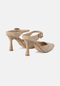 Zapatos de tacón alto de ante beige con punta afilada y tacón fino. Cuentan con una correa ajustable en el tobillo y un acento decorativo de joya.